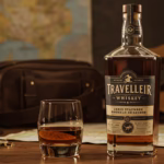 Cinematic shot of Traveller Whiskey bottle and bourbon glass on a rustic wooden table, with warm golden light, road map, leather bag, and guitar in the background.